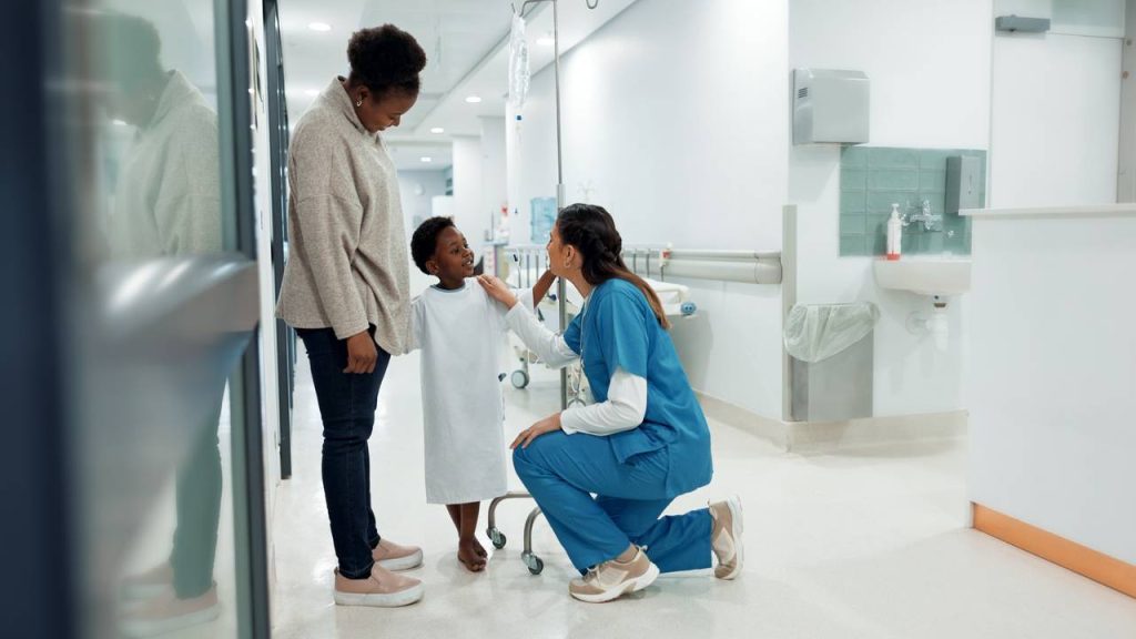 Nurse kneels in a hospital corridor speaking with a young patient while a parent stands beside them.
