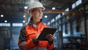 Construction supervisor wearing a hard hat and safety glasses, holding a tablet in an industrial warehouse.