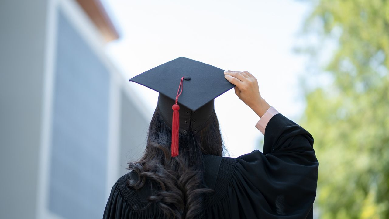 Graduate in a black gown seen from behind adjusting a mortarboard with a red tassel outdoors, with a blurred campus building and trees in the background.