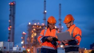 Two workers in high-visibility safety gear review a laptop and clipboard at an industrial site at dusk.
