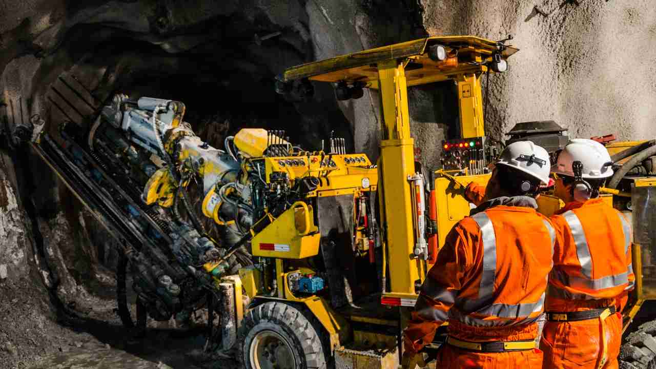 Two mining workers in orange safety gear standing beside underground drilling equipment in a mine tunnel